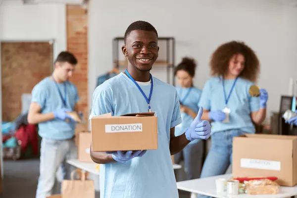 Portrait of african american young male volunteer in blue uniform, protective gloves smiling, showing thumbs up and holding cardboard box for Donation