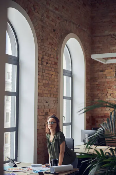 Дизайнер интерьера или архитектор в повседневной одежде с грязной прической, смотрящий в камеру во время работы над проектом, стоящий в ее офисе днем — стоковое фото