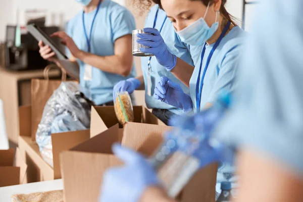 Närbild skott av upptagna frivilliga bär blå uniform, skyddsmasker och handskar sortera donerade livsmedel medan du arbetar i välgörenhet stiftelse — Stockfoto