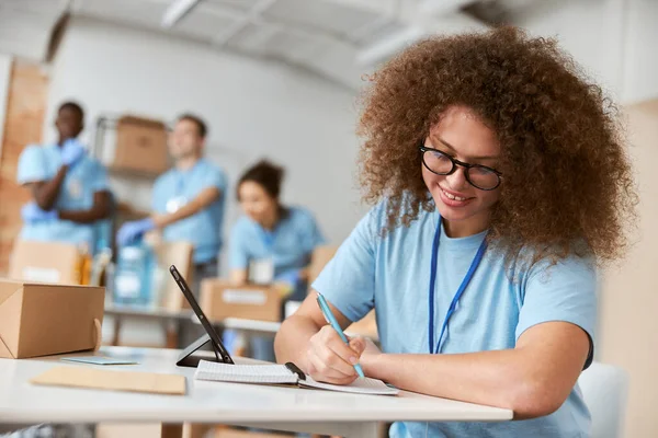 Ritratto di giovane volontaria in uniforme blu che prende appunti mentre lavora al progetto di donazione. Smistamento di squadra, imballaggio di oggetti in background — Foto Stock