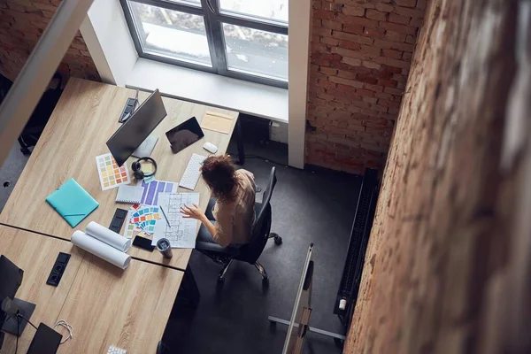 Вид сверху женщины-дизайнера, читающей синий принт, подбирающей цветовые образцы при работе над новым проектом дизайна интерьера в современном офисе — стоковое фото