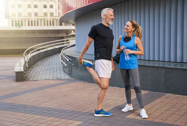 Orta yaşlı mutlu bir çift, spor kıyafetleri içinde gülümseyen bir adam ve bir kadın, dışarıda antremana hazır bir şekilde bekliyorlar.