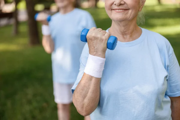 Leende äldre kvinna gör övningar med hantelträning med vän i grön park — Stockfoto