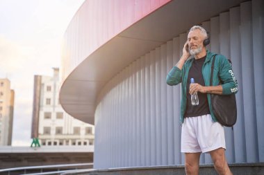 Spor giyim ve kulaklık takan, müzik dinleyen, dışarıda duran, tenis antrenmanına hazır, orta yaşlı, rahat bir adam.