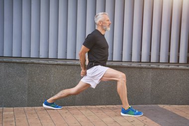 Spor kıyafetli, kendinden emin orta yaşlı bir adam bacaklarını esnetiyor, ısınıyor ve dışarıda egzersiz yapıyor.
