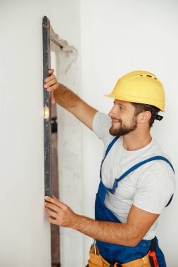 Mutlu tamirci, üniformalı müteahhit balon seviyesini kullanarak duvarı ölçerken gülümsüyor.