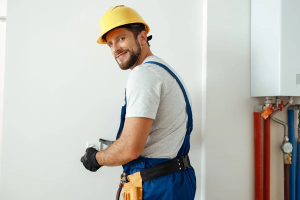 Selbstbewusster Elektriker mit Mütze und Schutzhandschuhen lächelt in die Kamera, während er Steckdose in einem Raum installiert — Stockfoto