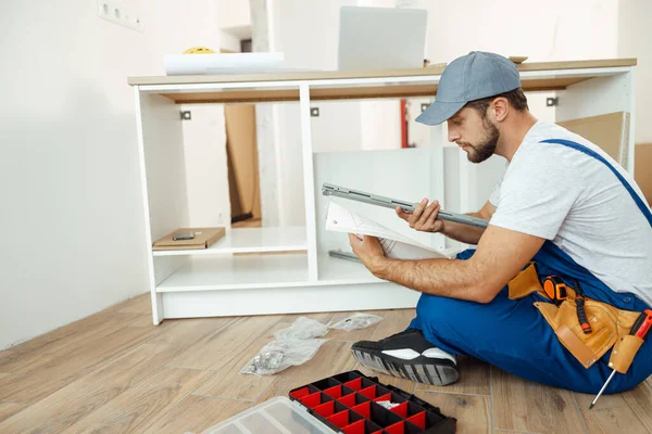 Drukke klusjesman in overalls zittend op de vloer en leeshandleiding bij het monteren van keukenkastje in appartement — Stockfoto