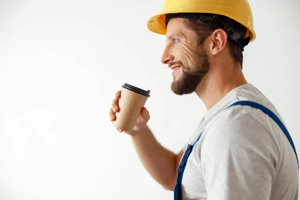 Samping melihat potret tukang yang tersenyum dengan seragam dan topi keras sedang minum kopi selama istirahat sambil berdiri di dalam ruangan — Stok Foto