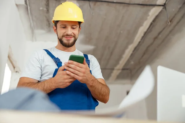 Focused repairman in workwear and hardhat smiling while using smartphone, standing indoors during renovation work — Φωτογραφία Αρχείου