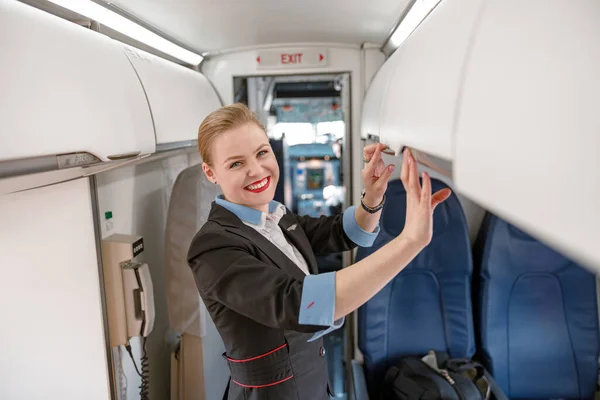 Cheerful flight attendant checking overhead shelf in airplane — Stock fotografie