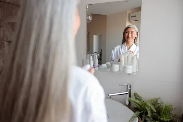 Wanita cantik berdiri di depan cermin di kamar mandi selama perawatan kulit pagi — Stok Foto