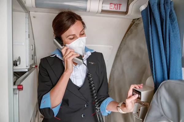 Žena letuška v masce mluví na telefonu v letadle — Stock fotografie