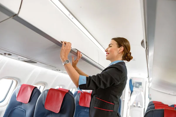 Alegre azafata cerrando la papelera de equipaje en el avión —  Fotos de Stock