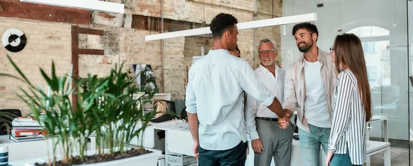 Dos hombres estrechando la mano y sonriendo mientras están de pie con sus compañeros de trabajo en la oficina creativa — Foto de Stock