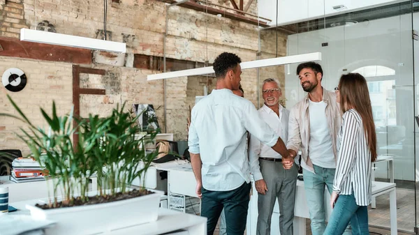 Due uomini che si stringono la mano e sorridono in piedi con i colleghi nell'ufficio creativo — Foto Stock