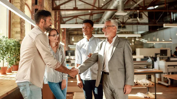 Trevligt att träffas. Två glada män skakar hand och ler när de står med arbetskamrater på kreativa kontor. — Stockfoto