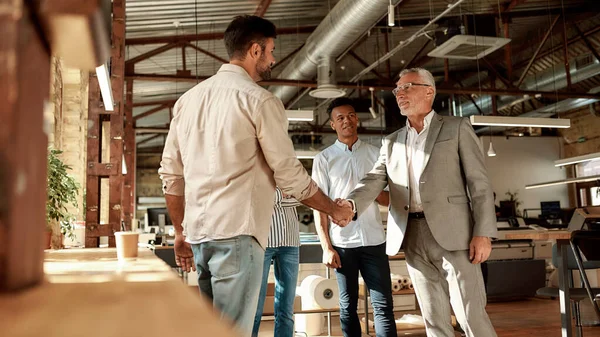 Bienvenidos a nuestro equipo. Dos hombres alegres estrechando la mano y sonriendo mientras están de pie con sus compañeros de trabajo en la oficina moderna — Foto de Stock