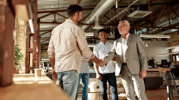 Welkom in ons team. Twee vrolijke mannen schudden handen en glimlachen terwijl ze staan met collega 's in het moderne kantoor — Stockfoto