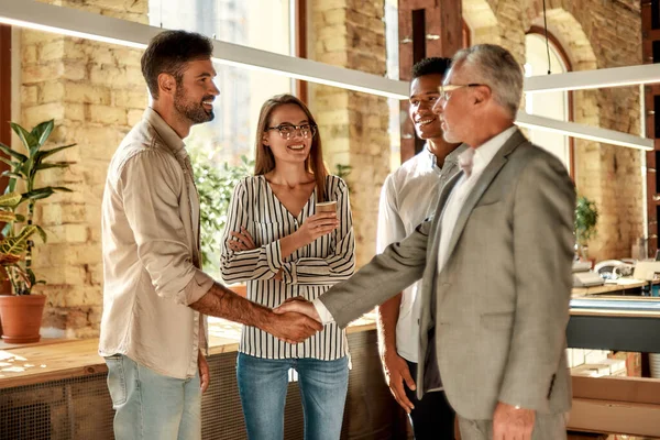 Två affärsmän skakar hand och ler när de står med medarbetare på det kreativa kontoret. — Stockfoto