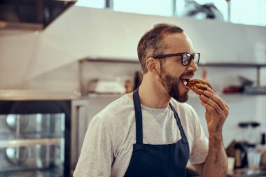 Önlüklü sakallı adam kafede hamur işi yiyor.