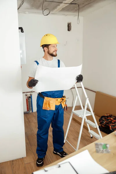 Full length shot of thoughtful contractor in overalls and hard hat looking away while studying blueprint, standing in new apartment — Stock Photo, Image