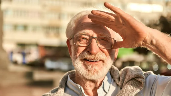 Retrato de homem idoso barbudo feliz e bonito em óculos olhando para a câmera e sorrindo enquanto passa o tempo ao ar livre em um dia ensolarado — Fotografia de Stock
