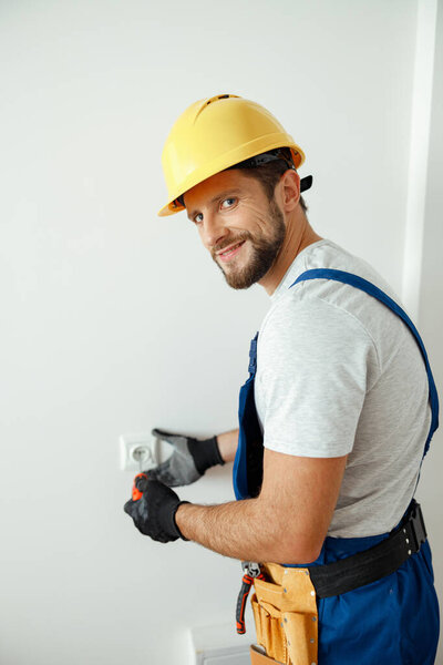 Happy male worker, professional electrician in hard hat and protective gloves smiling at camera while replacing electrical socket in a room. Manual work, maintenance concept