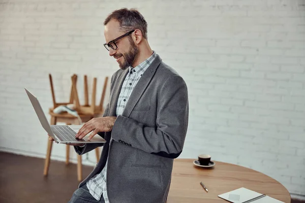 ポータブルコンピュータを持ちながら テーブルの端に座って笑顔の楽しい男性 — ストック写真