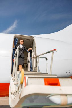 Neşeli bir uçuş görevlisi, özel bir jetin kapısında dururken merdivenlere bakıyor ve gülümsüyor.