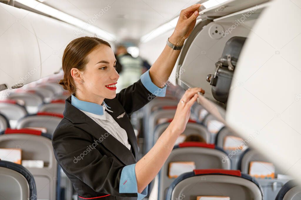 Cheerful woman flight attendant looking at travel suitcase in overhead baggage bin and smiling while standing in aircraft passenger salon
