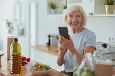 Mutfakta cep telefonu kullanan neşeli emekli kadın.