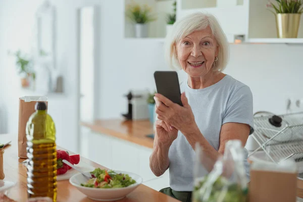 Wanita pensiunan yang ceria menggunakan ponsel di dapur — Stok Foto