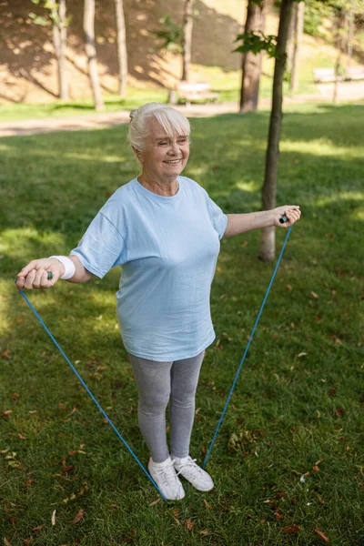 Glückliche Seniorin in Sportkleidung turnt mit Springseil im grünen Park — Stockfoto