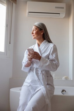 Bornozlu güzel bir Asyalı kadın banyoda akıllı telefon tutuyor.