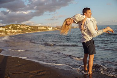 Güçlü bir adam kız arkadaşını omzunda tutarken gelen dalgaların yanında kumların üzerinde duruyor.