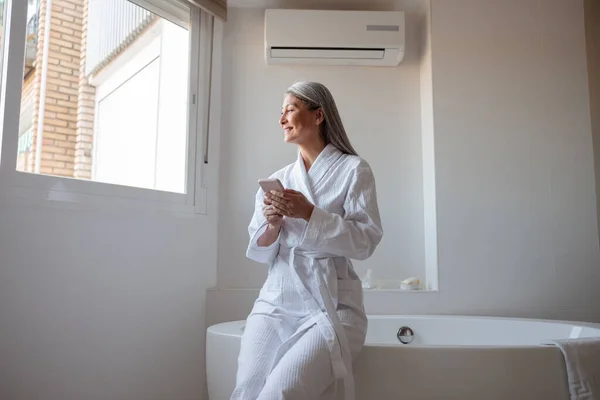 Souriant dame asiatique dans la salle de bain assis sur la baignoire et tapant sur smartphone — Photo