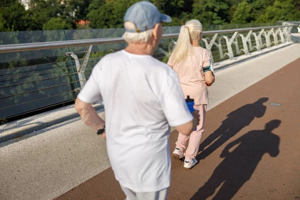 Par de pensionistas ativos corre juntos ao longo da ponte pedonal na vista traseira do dia de verão — Fotografia de Stock