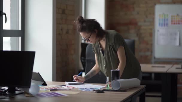 Lavori in corso. Giovane designer donna in abbigliamento casual cercando occupato, prendendo appunti e schizzi mentre in piedi sul suo posto di lavoro in ufficio — Video Stock