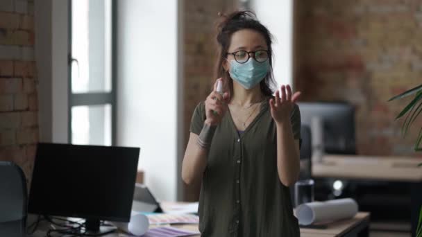 Preventive measures. Young woman, creative designer wearing protective mask spraying sanitizer gel to clean her hands while standing in her office — Stock Video