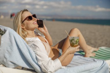 Güverte sandalyesinde uzanmış gülümseyen kadın, kumsalda kokteyl içiyordu.