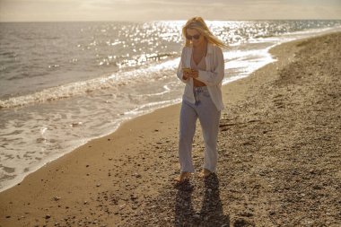 Gün batımında sahilde yürürken akıllı telefondan mesaj yazan barışçıl bir kadın.