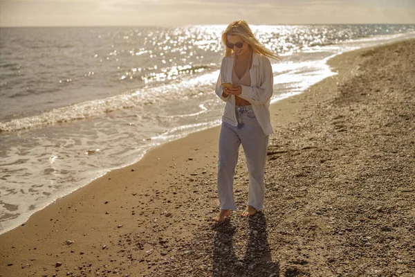 Gün batımında sahilde yürürken akıllı telefondan mesaj yazan barışçıl bir kadın.