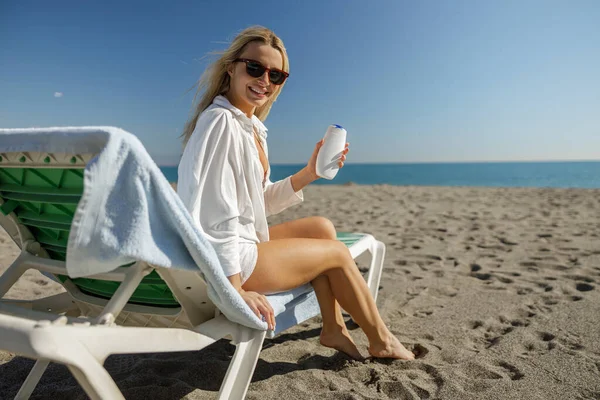 Güneş gözlüklü sarışın kadın kameraya gülümsüyor, plajda güneşlenirken krem sürüyor.