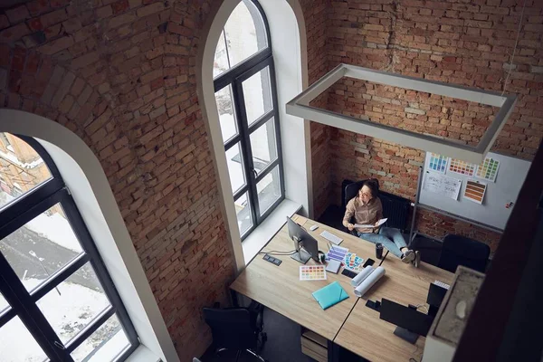Masada oturup modern ofiste iç tasarım projesi üzerinde çalışırken pencereye bakan yaratıcı kadının yüksek açılı görüntüsü