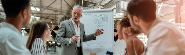 Homme d'affaires mature confiant pointant du doigt le tableau blanc et expliquant quelque chose à ses jeunes collègues lors d'une réunion au bureau — Photo