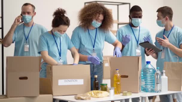 Team of diverse young volunteers in protective gloves with masks while sorting, packing foodstuff in cardboard box, working together on donation project indoors — Vídeos de Stock