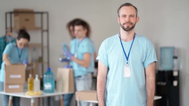 Smiling caucasian male volunteer in blue uniform and protective gloves standing with arms crossed. Team sorting, packing items in cardboard boxes — Vídeos de Stock