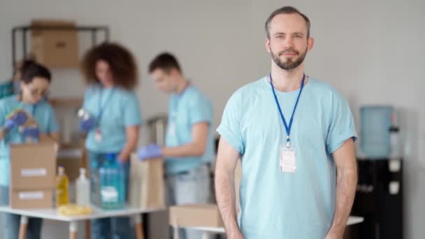 Caucasian male volunteer in blue uniform showing heart love sign, looking at camera. Team sorting, packing items in cardboard boxes — Vídeo de stock