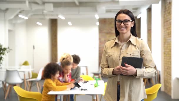 Cheerful young female teacher in glasses smiling at camera, standing in a classroom. Kids sitting at the table, studying in elementary school — Video Stock